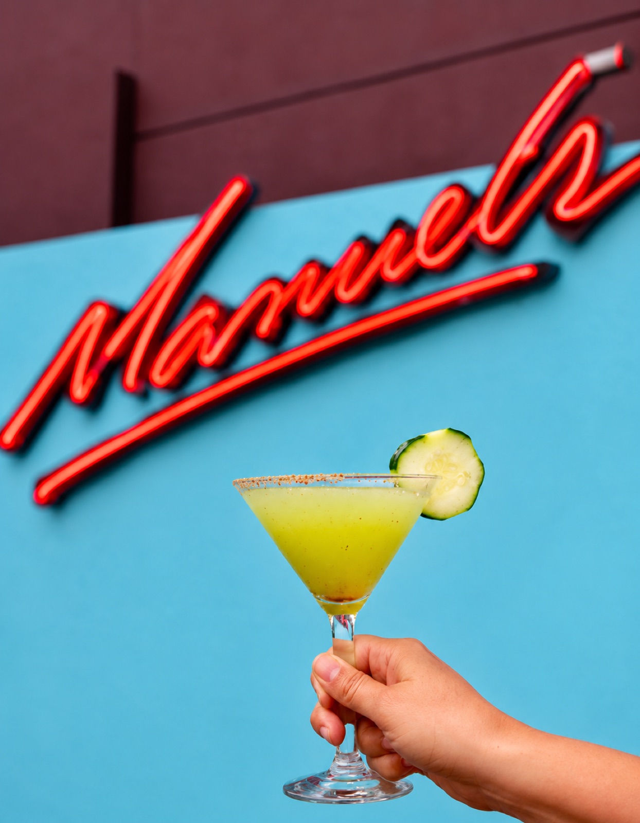 A hand raising a cucumber-lime martini with a cucumber garnish in front of Manuel's red neon sign
